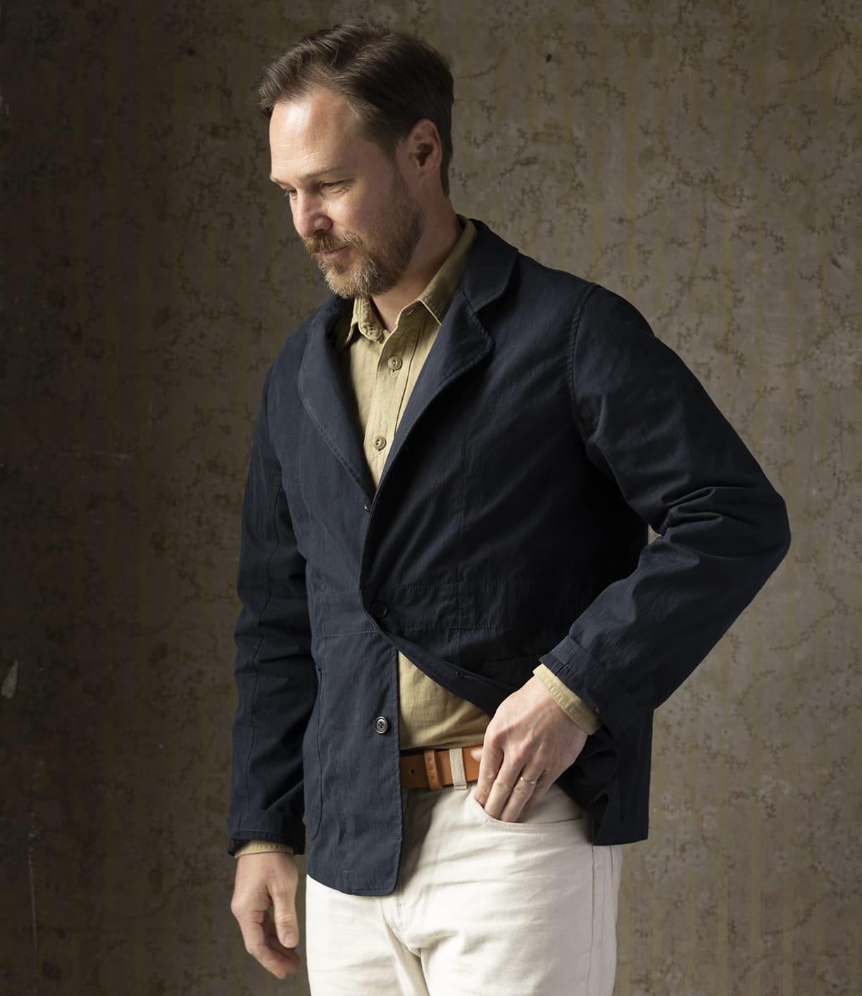 A man with short brown hair and a beard stands against a textured wall, wearing the Best Made Co Possibilities Jacket Navy with white pants and a tan belt, looking down thoughtfully as he adjusts his jacket.