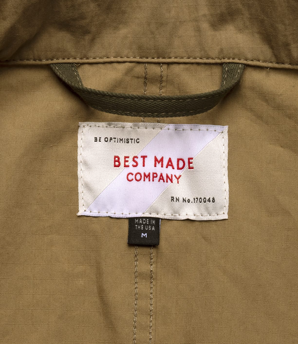 A close-up of a tan fabric label reads BEST MADE CO in red, with a black tag stating MADE IN THE USA M, highlighting American quality—just like the Possibilities Jacket Tan from Best Made Co.