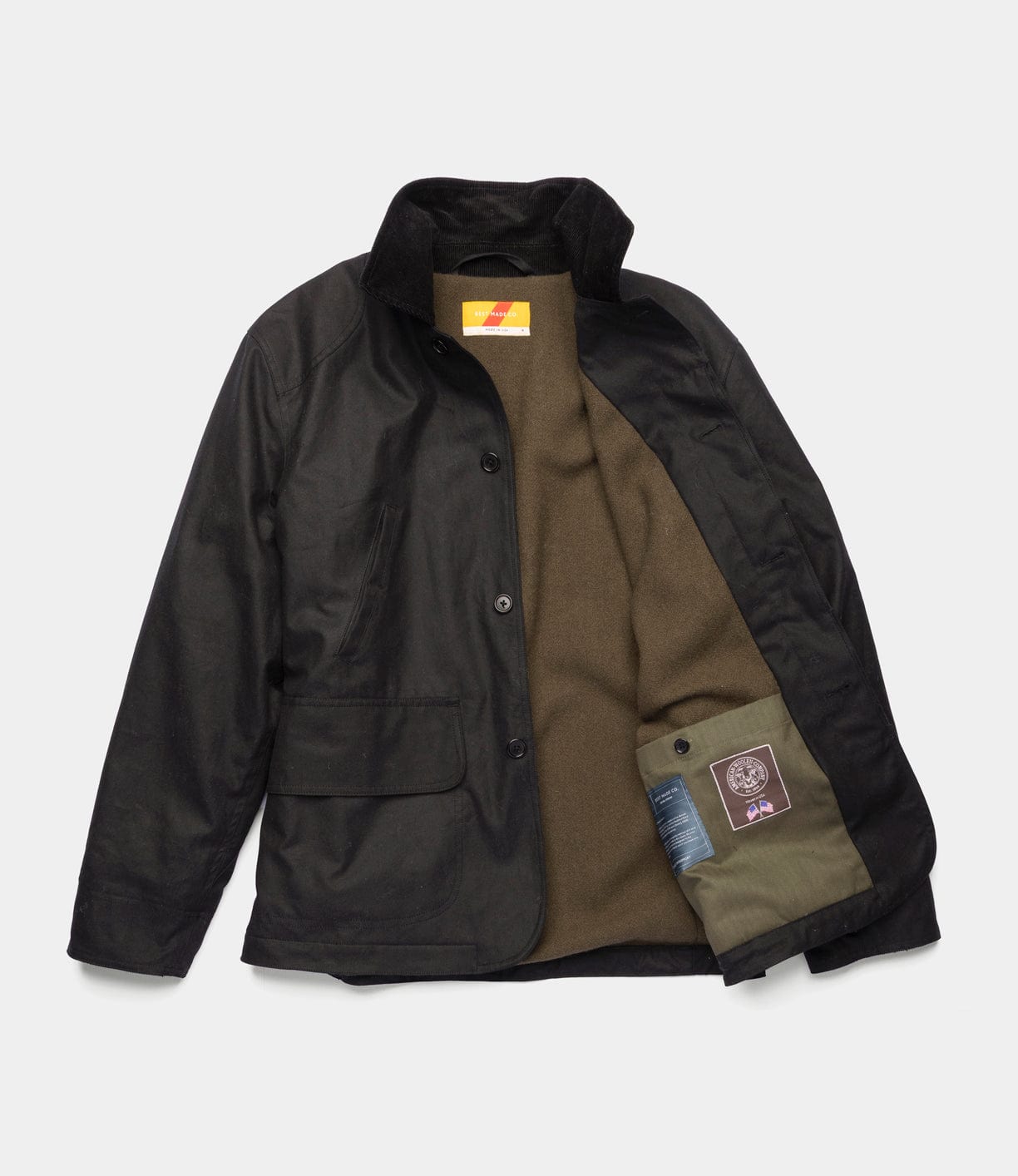 The Best Made Co. Wool-Lined Waxed Field Jacket Black, shown partially open on a white background, features a corduroy collar, Wyoming wool lining, interior label, and a green inner pocket with a patch and blue tag.