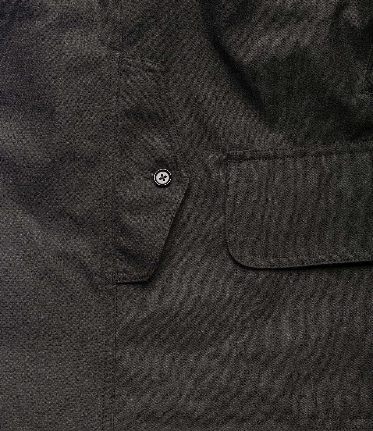 Close-up of the Best Made Co. Wool-Lined Waxed Field Jacket Black shows a smooth, slightly matte British Millerain Staywax fabric with a water-resistant finish, flap pocket, visible stitching, and button detail on the left side.