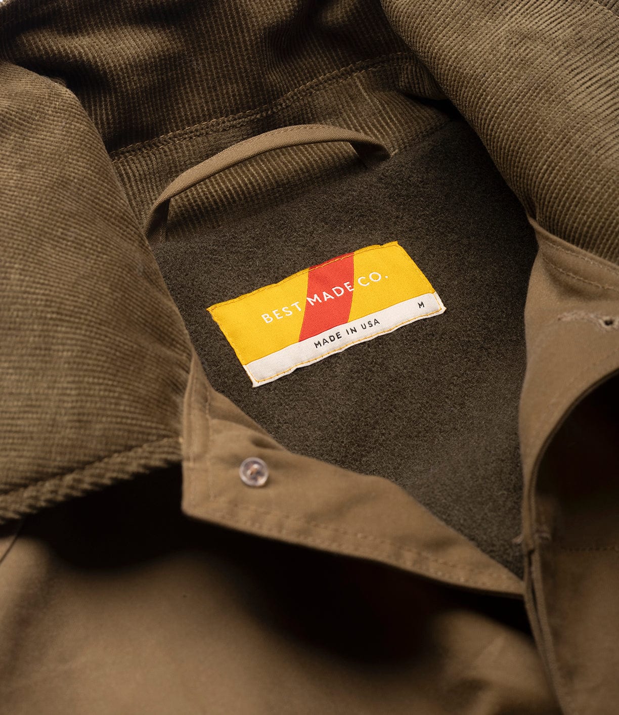 Close-up of the Wool-Lined Waxed Field Jacket Tan collar by Best Made Co., featuring a yellow and white label marked Made in USA, size M. The image highlights the tan waxed cotton texture, detailed stitching, and weather-resistant finish.