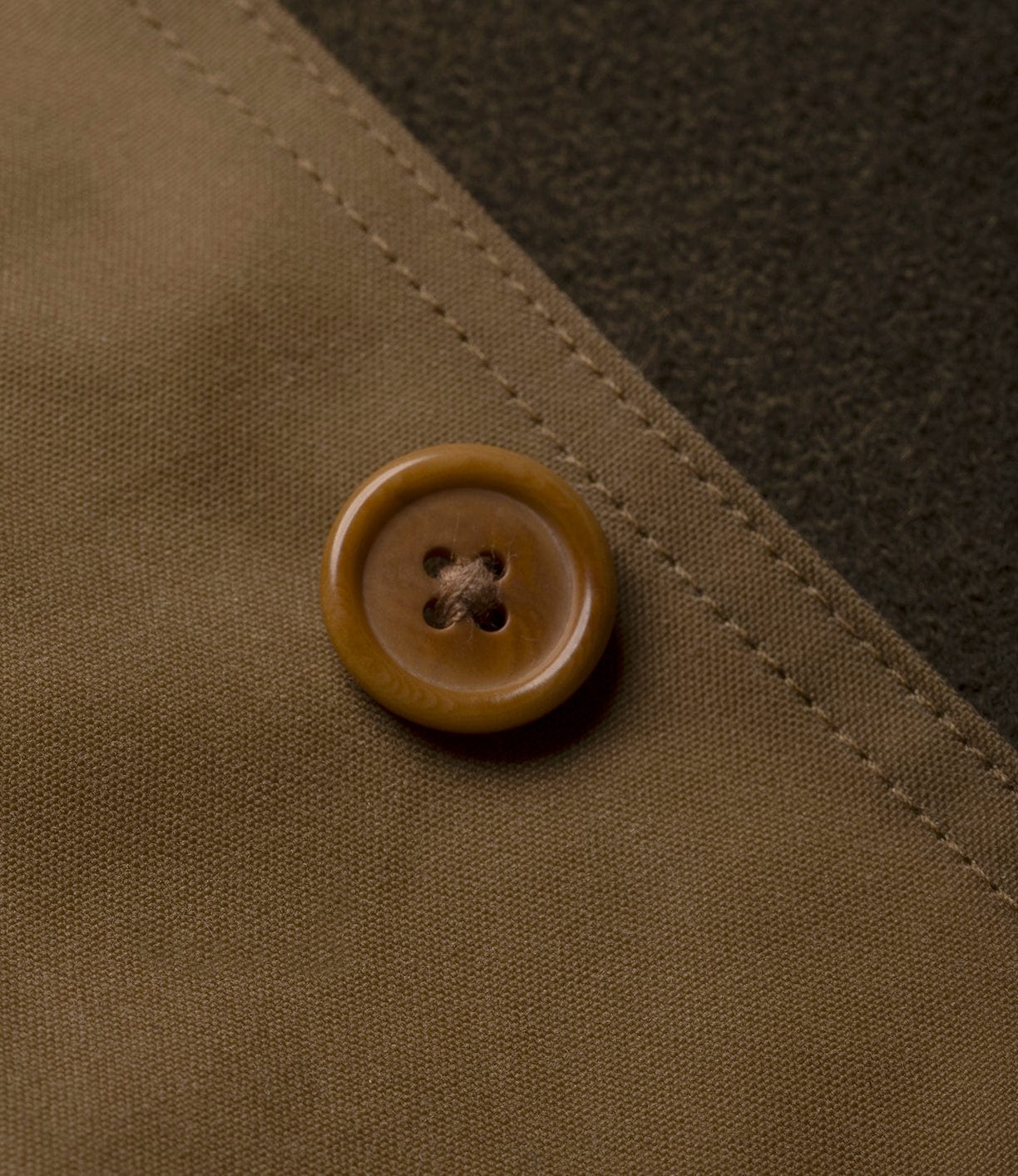 Close-up of a brown, four-hole button on light tan fabric with visible stitching—detailing the Wool-Lined Waxed Field Jacket Tan by Best Made Co. A darker textured material in the background adds visual contrast.