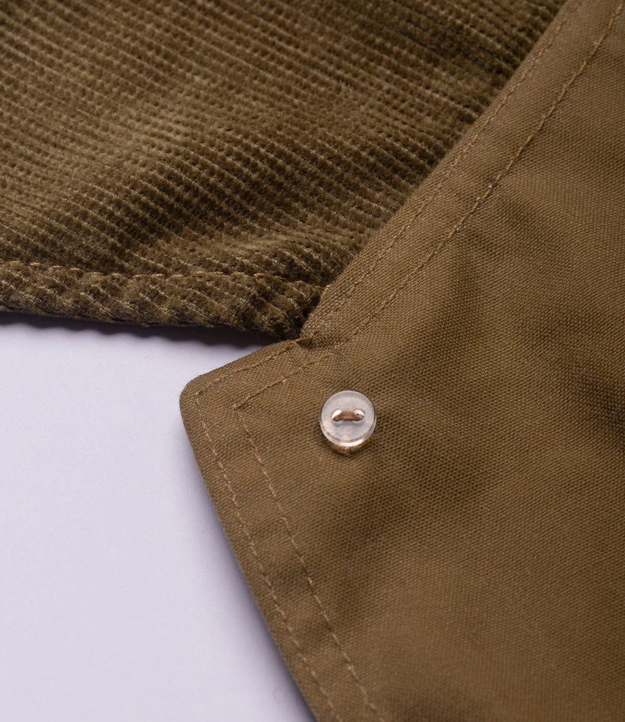 Close-up of the Best Made Co. Wool-Lined Waxed Field Jacket in Tan, highlighting brown corduroy and smooth fabric, a clear button, and white stitching—signature details against a light background.