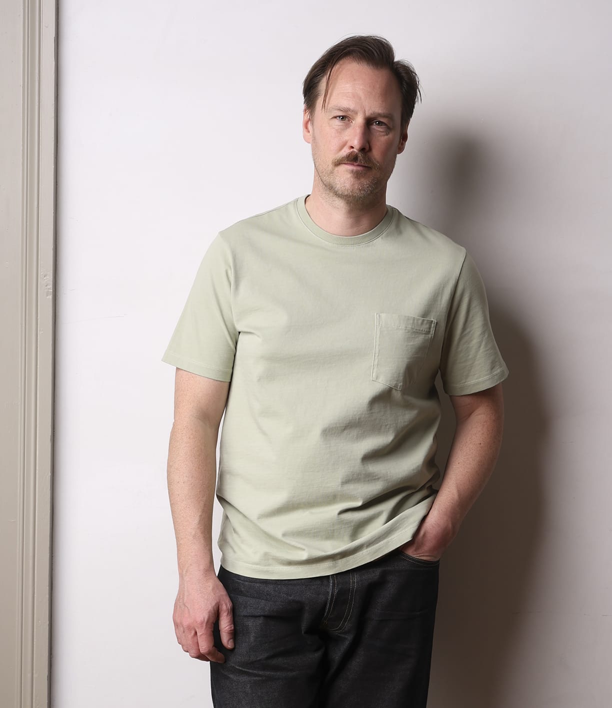A man with light brown hair and a mustache stands against a plain light wall, wearing the Best Made Co Standard Pocket T-Shirt in Haze and dark jeans, one hand in his pocket, with a neutral expression.