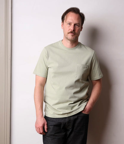 A man with light brown hair and a mustache stands against a plain wall, wearing the Best Made Co. Standard Pocket T-Shirt in Haze—a pale green garment-dyed tee with a chest pocket—and dark jeans, one hand in his pocket and a neutral expression.