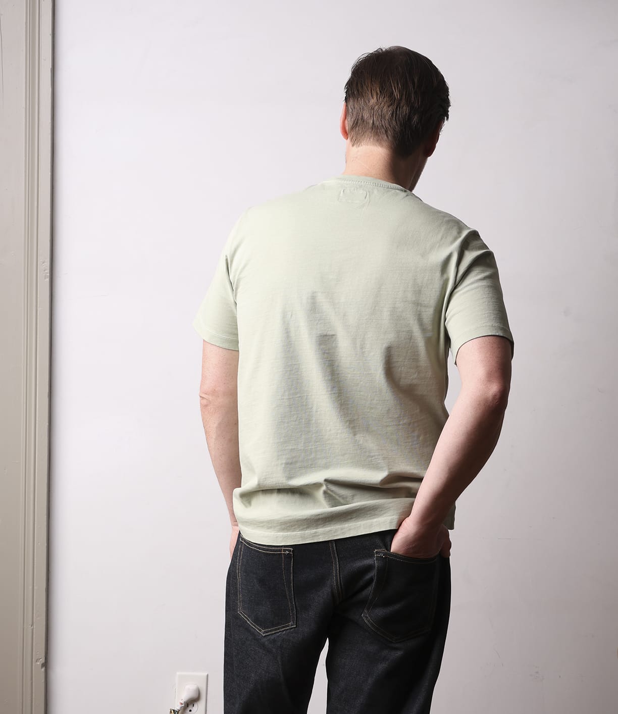 A person with short hair stands facing away from the camera, wearing the Best Made Co Standard Pocket T-Shirt Haze in light green and dark jeans, with one hand in their back pocket against a plain white wall.