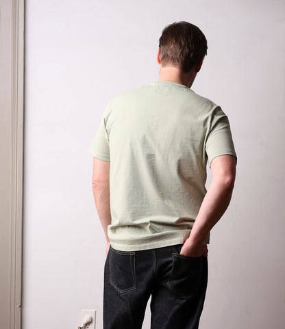 A person stands against a light wall wearing the Best Made Co. Standard Pocket T-Shirt Haze—crafted from durable Peruvian tangüis cotton—with dark jeans. One hand is in their pocket, the other on their lower back, highlighting this everyday staple.