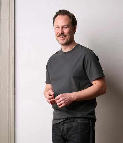 A man with light brown hair and a mustache stands smiling against a white wall, wearing the Best Made Co. Standard T-Shirt in Faded Black and dark jeans, with his hands loosely clasped in front of him.