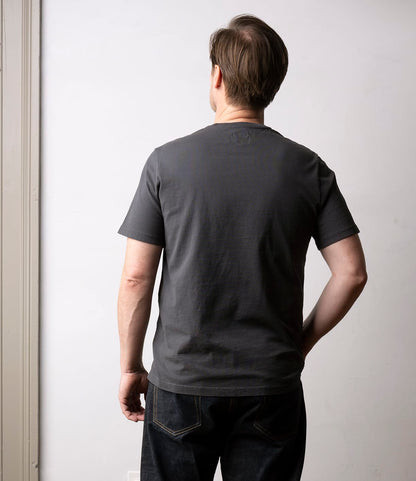 A man with short brown hair faces away from the camera, wearing Best Made Co.’s Standard T-Shirt in Faded Black with dark jeans, standing against a plain light-colored wall.