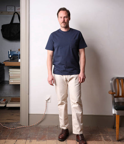 A man with short hair and a beard stands indoors on a wooden floor, wearing the Best Made Co. Standard Pocket T-Shirt in navy with beige pants and brown shoes. A chair and a shelf holding books and a sewing machine are visible in the background.