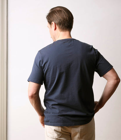 A man faces away from the camera, wearing the Best Made Co. Standard Pocket T-Shirt in navy and light-colored pants, with his hands on his hips against a plain light background.