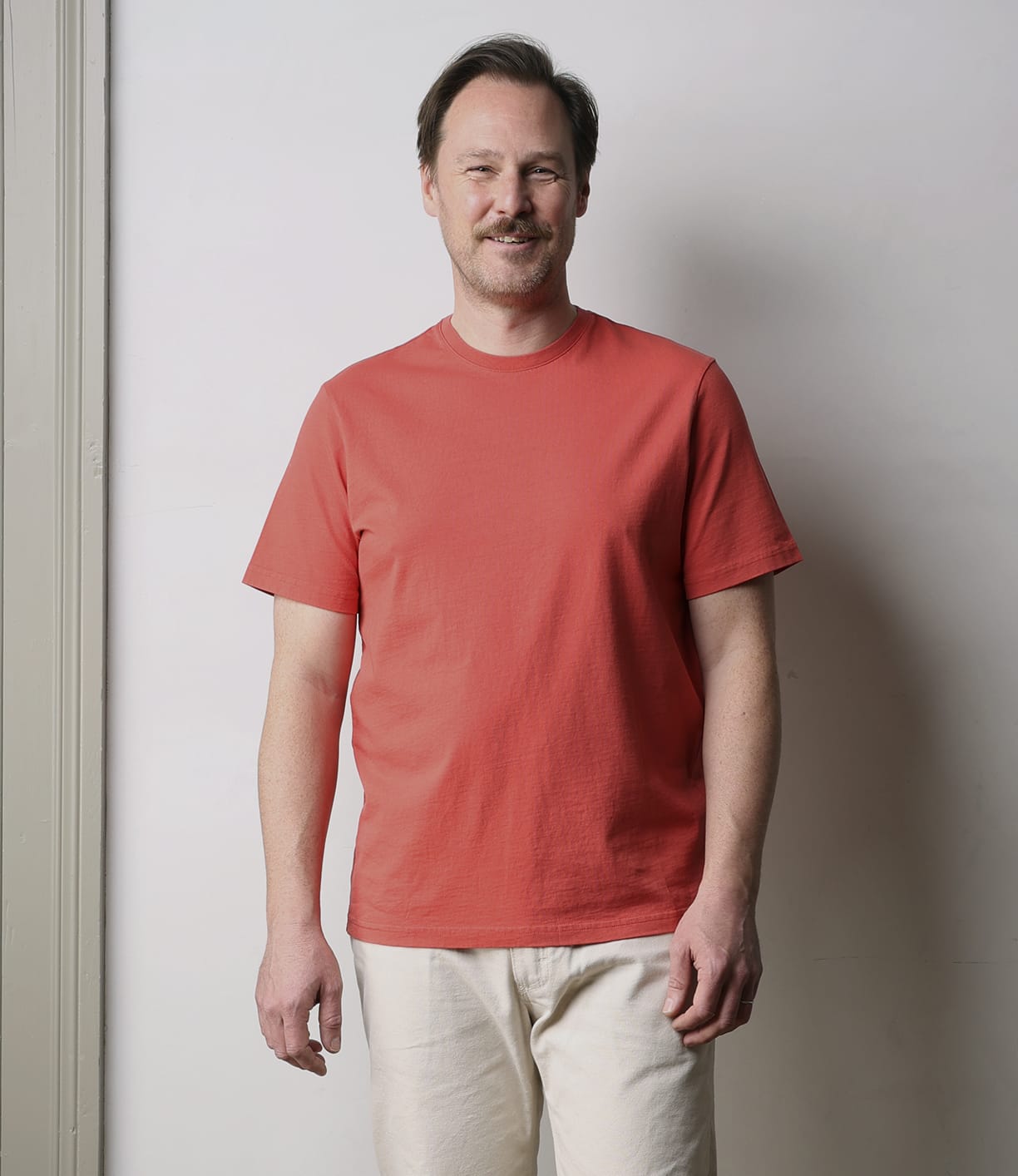 A man with short brown hair and a mustache smiles against a plain light wall, wearing Best Made Co's Standard T-Shirt in Faded Red and light beige pants for a durable everyday staple look.