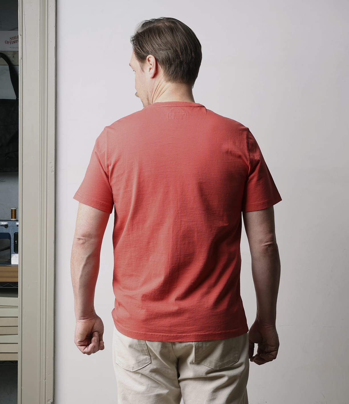 A man with short brown hair stands indoors facing away from the camera, wearing a Best Made Co Standard T-Shirt in Faded Red and beige shorts in front of a plain light gray wall.