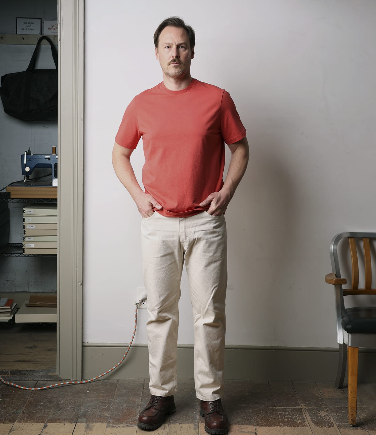 A man stands indoors against a plain wall, wearing the Best Made Co Standard T-Shirt in Faded Red, beige pants, and brown shoes. His hands are in his pockets. A chair, books, and a sewing machine are visible in the background.