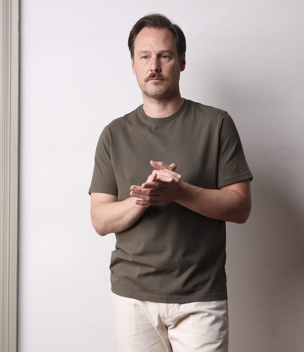 A man with light brown hair and a mustache stands by a light wall, wearing Best Made Co’s Standard T-Shirt Field Drab in soft Peruvian tangüis cotton and light beige pants, looking slightly to the side with his hands clasped.