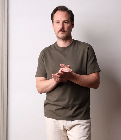 A man with light brown hair and a mustache wears the Best Made Co. Standard T-Shirt Field Drab, made from Peruvian tangüis cotton, and light beige pants, standing against a plain white wall with his hands clasped, looking forward.