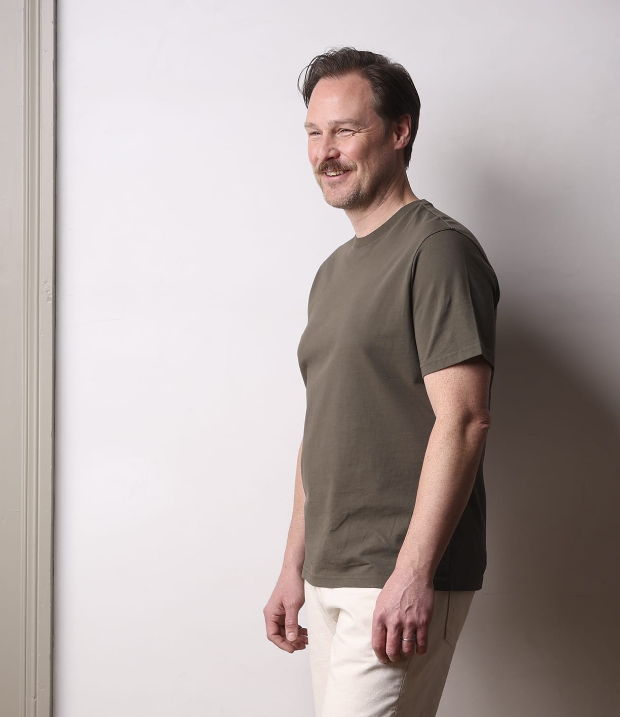 A man with short hair and a mustache stands sideways, smiling, wearing the Best Made Co Standard T-Shirt in Field Drab and light-colored pants against a plain light wall.