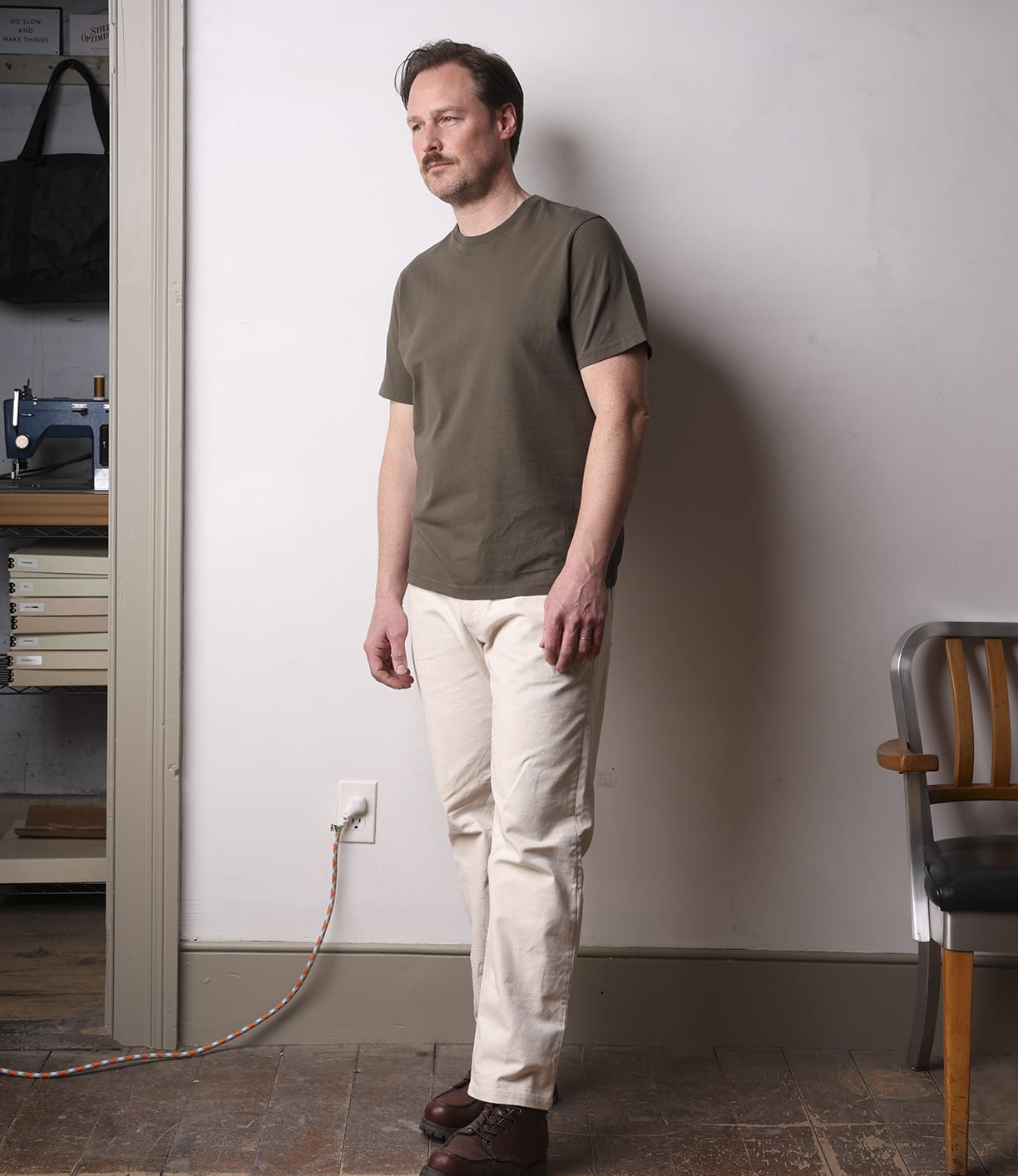 A man stands indoors by a light wall, wearing the Best Made Co Standard T-Shirt Field Drab, light beige pants, and brown shoes. A wooden chair and a shelf with books and equipment are partially visible in the background.