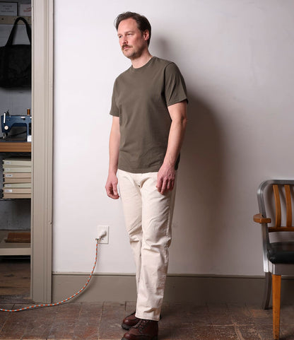 A man stands indoors against a white wall, wearing the Best Made Co. Standard T-Shirt in Field Drab, light-colored pants, and brown shoes. Next to him is a wooden chair and a nearby plugged-in power cord.
