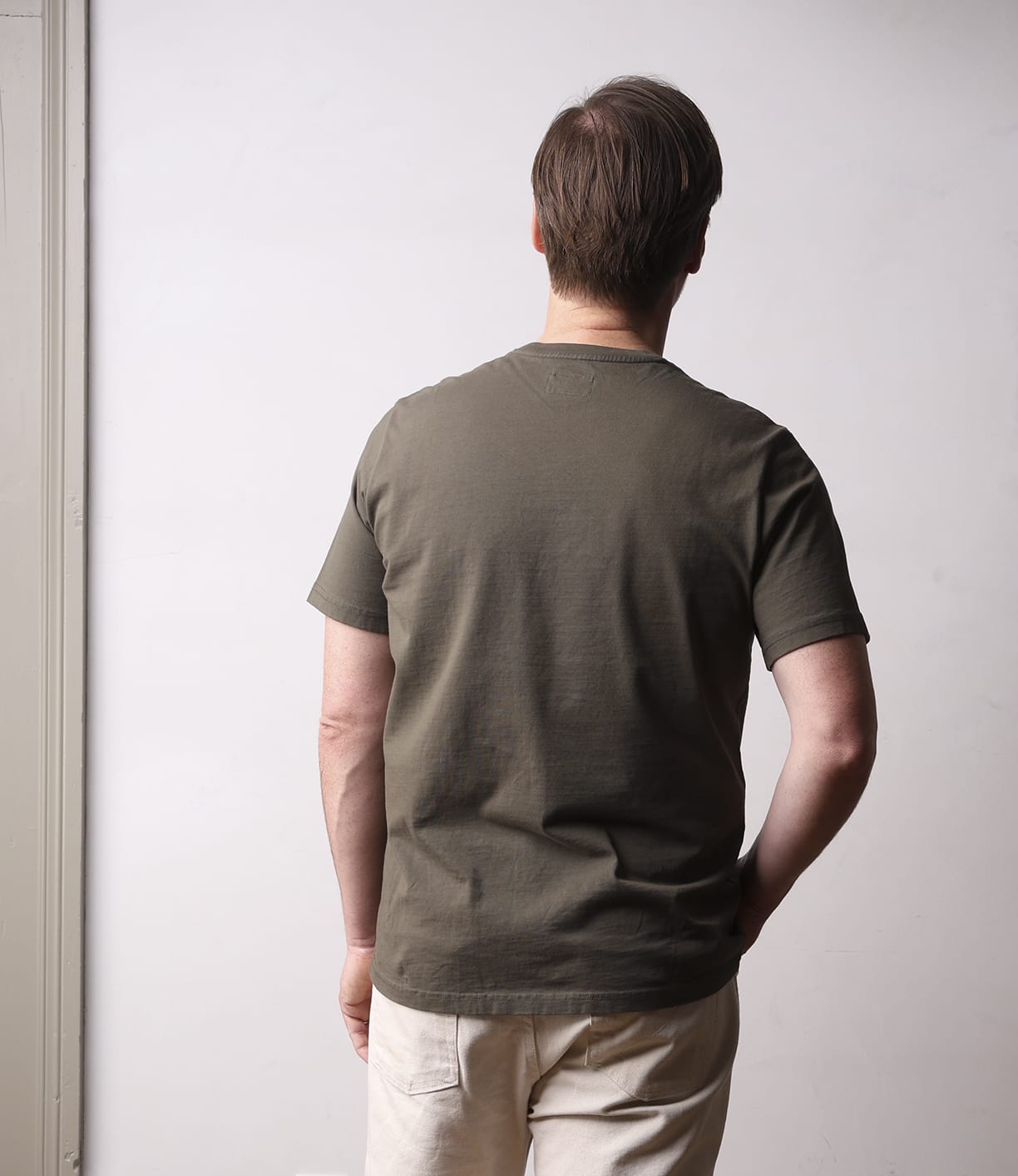 A man with short brown hair faces away from the camera, wearing the Best Made Co Standard T-Shirt Field Drab and light pants, standing against a plain light-colored wall.