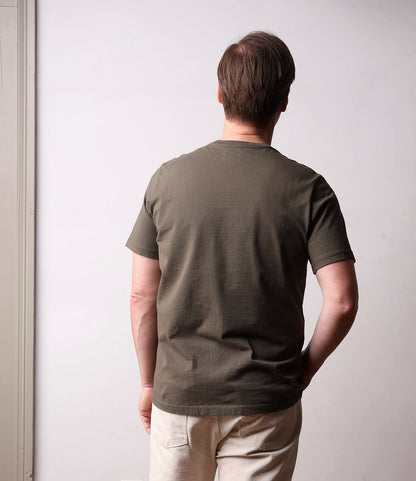 A person with short brown hair, wearing the Best Made Co. Standard T-Shirt in Field Drab and light pants, stands facing away from the camera against a plain light-colored wall.