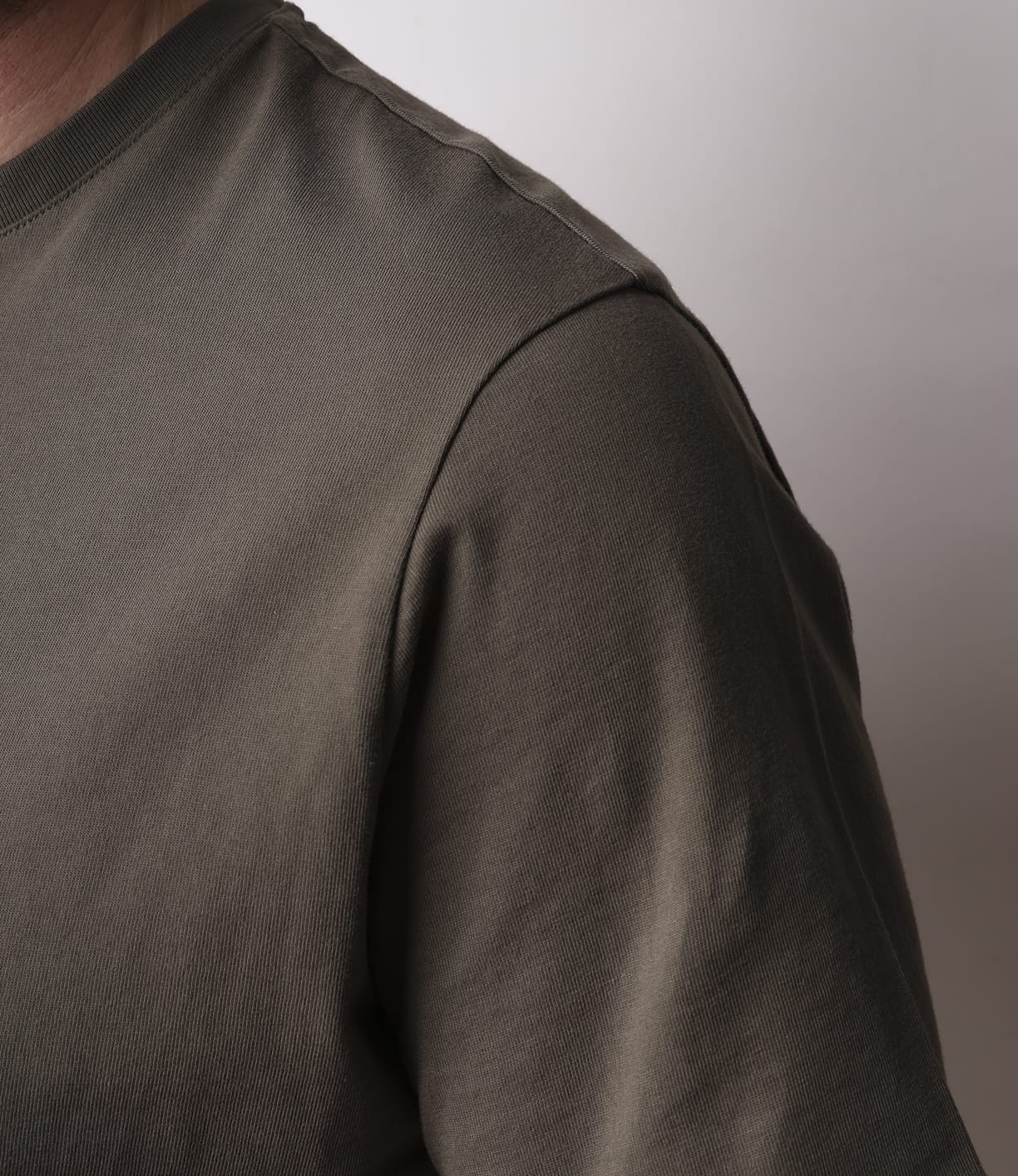 A close-up of a person in the Best Made Co Standard T-Shirt Field Drab, made from soft Peruvian tangüis cotton, highlights the olive green shoulder, sleeve, and upper chest. The plain light background keeps the face unseen.