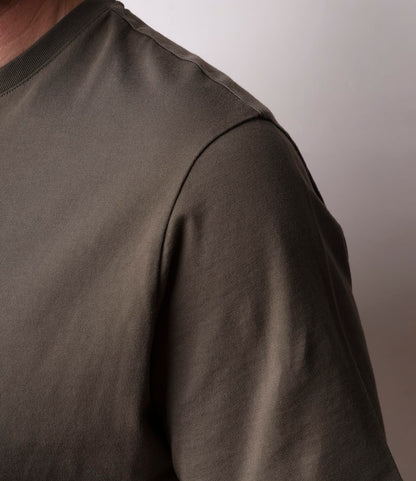 Close-up of a person wearing the Best Made Co. Standard T-Shirt Field Drab, featuring soft Peruvian tangüis cotton in olive green. Only the shoulder, upper chest, and sleeve are visible against a plain light background; head and lower body are out of frame.