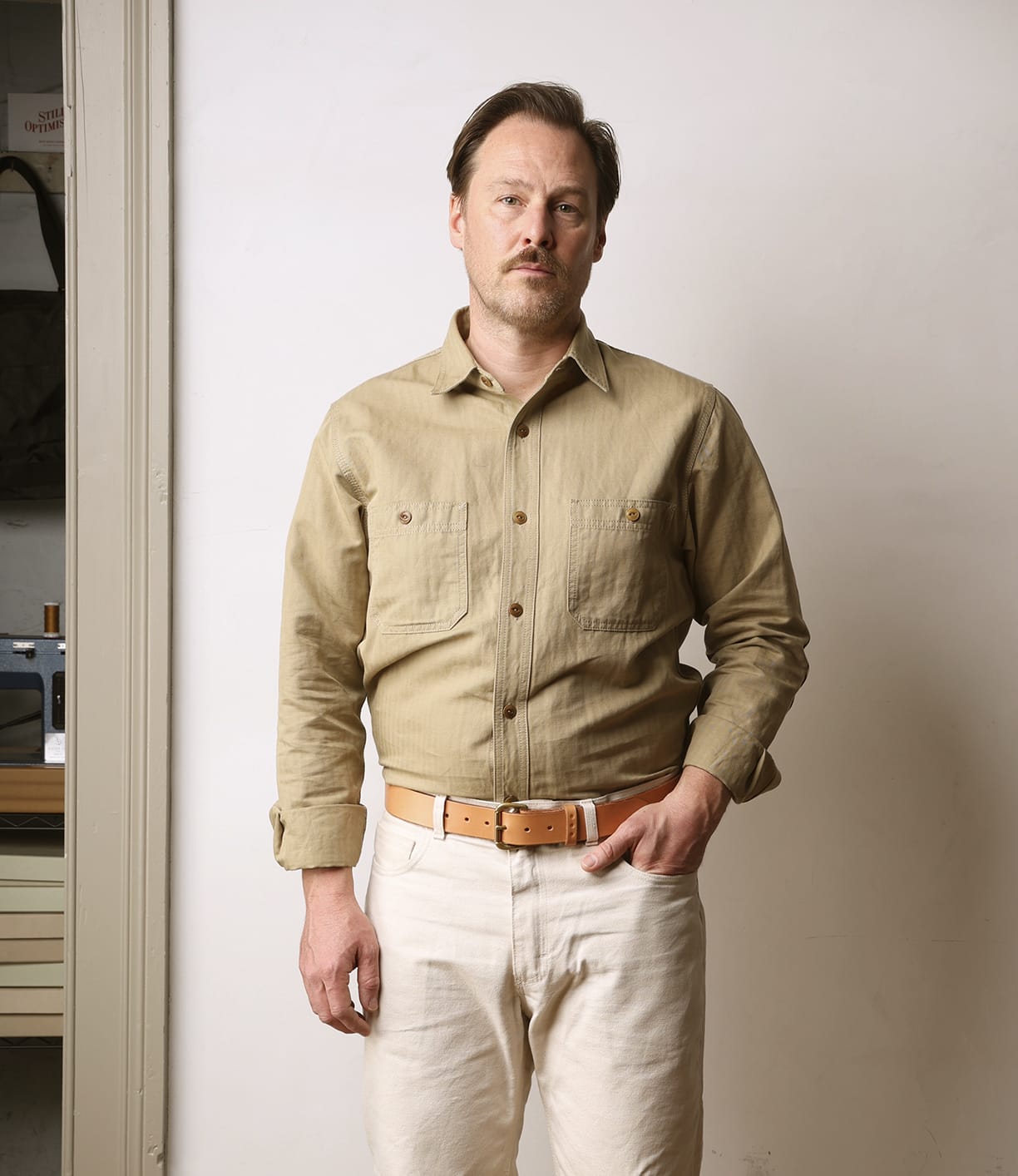 A man with short hair and a mustache stands against a plain wall, wearing the Best Made Co. Cotton-Linen Workshirt in Tan, paired with light pants and a beige belt; one hand is in his pocket and he has a neutral expression.