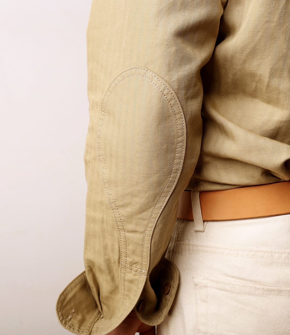 Close-up of a person wearing the Best Made Co. Cotton-Linen Workshirt in Tan, featuring an oval elbow patch and paired with cream pants and a tan belt. The sleeve is slightly wrinkled. Made in Portugal.
