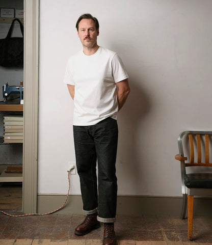 A man with short brown hair and a mustache stands indoors against a white wall, wearing the Best Made Co. Standard T-Shirt Undyed, cuffed black jeans, and brown boots. His hands are behind his back; nearby are a chair and sewing machine.