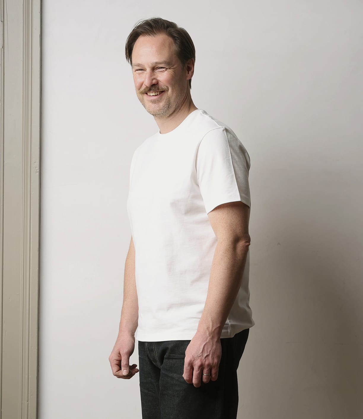 A man with light brown hair and a mustache, wearing the Best Made Co Standard T-Shirt Undyed—crafted from durable Peruvian tangüis cotton—and dark jeans, stands smiling against a plain, light-colored wall.