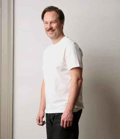 A man with light brown hair and a mustache smiles in a Best Made Co. Standard T-Shirt Undyed and black jeans, standing by a light-colored wall and door frame.