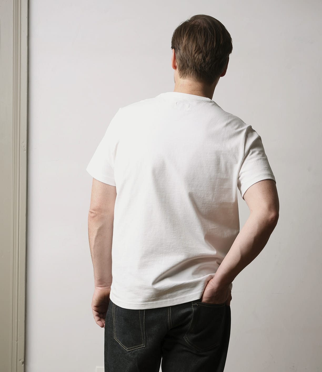 A person with short brown hair, wearing the Best Made Co Standard T-Shirt Undyed and dark jeans, stands facing away from the camera against a light-colored wall, one hand on their hip.