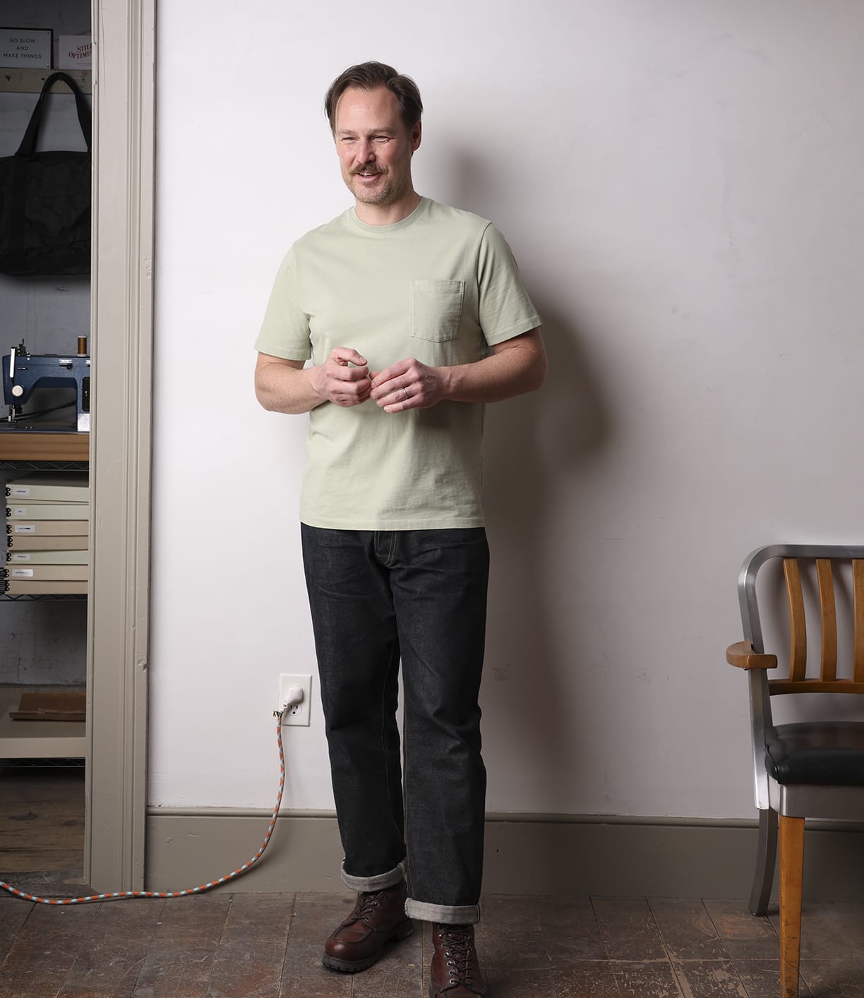 A man with short hair wears the Best Made Co Standard Pocket T-Shirt Haze, paired with dark jeans and brown boots. He stands indoors near a wooden chair and wall outlet, smiling slightly with his hands loosely clasped.