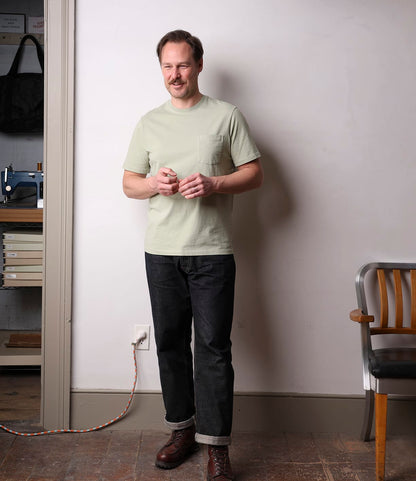 A man wearing the Best Made Co. Standard Pocket T-Shirt Haze stands indoors near a wooden chair, smiling with his hands together. A sewing machine and a coiled extension cord are visible in the background.