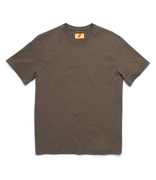 A man stands indoors by a light wall, wearing the Best Made Co Standard T-Shirt Field Drab, light beige pants, and brown shoes. A wooden chair and a shelf with books and equipment are partially visible in the background.