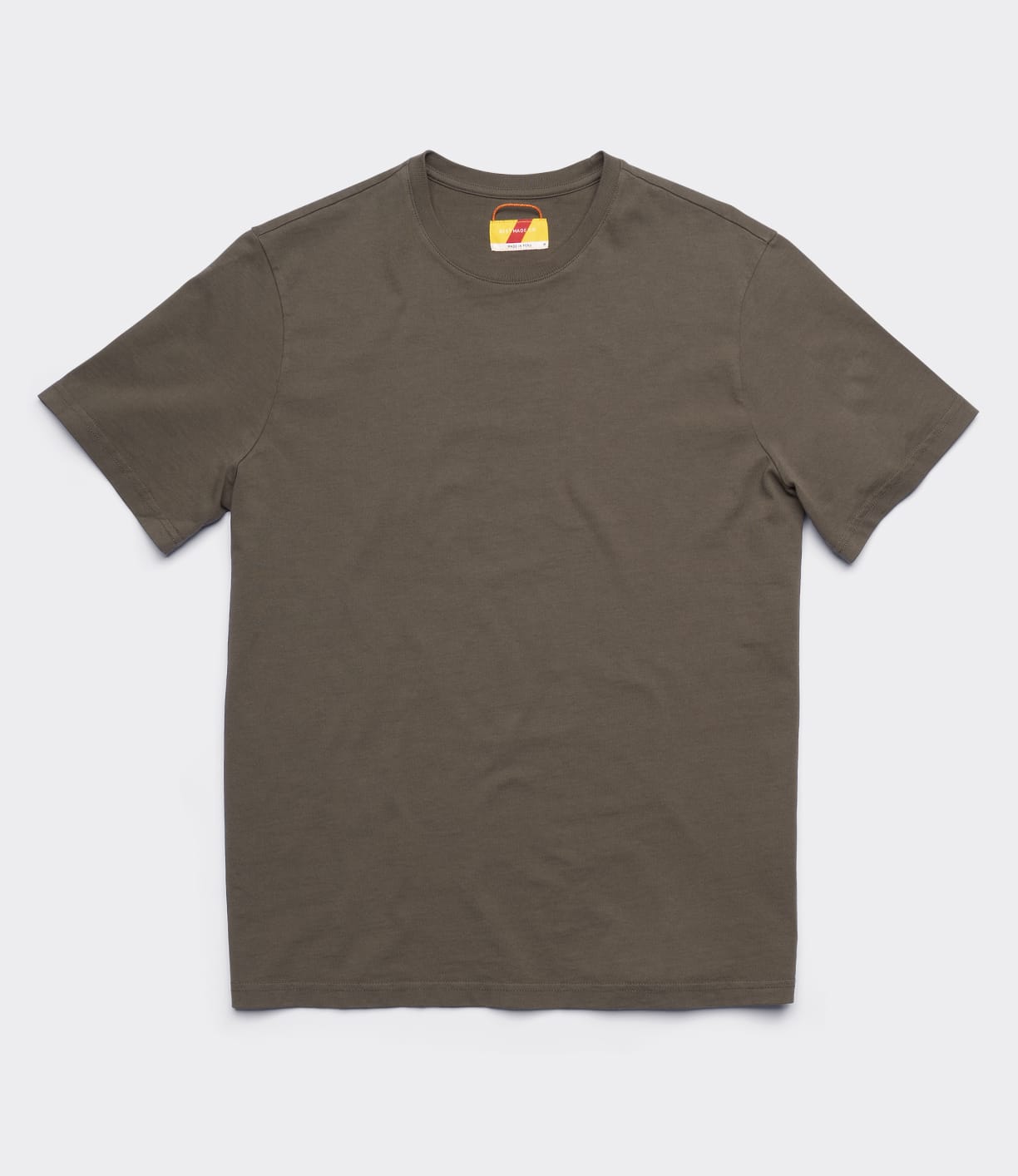 The Best Made Co Standard T-Shirt Field Drab, a garment-dyed, short-sleeve, crew neck tee in solid brownish-gray, is laid flat on a light gray background. The inside neck tag with a yellow and red design is visible.