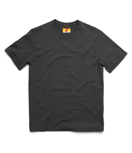 A man indoors stands on a wooden floor near a wall socket, wearing the Best Made Co Standard T-Shirt in Faded Black with dark jeans and brown boots. One hand in his pocket, he gestures; a chair and sewing machine complete this everyday staple look.