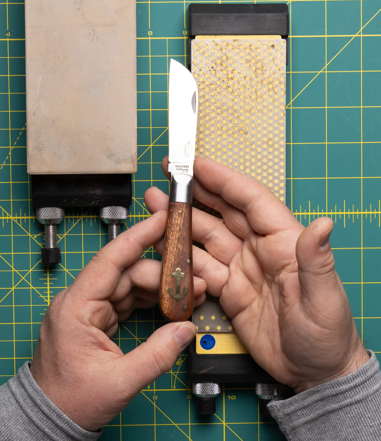 A person holds the Best Made Co. Anchor Knife, featuring a wooden handle and sheepsfoot blade, over sharpening stones on a green mat, preparing to sharpen it.