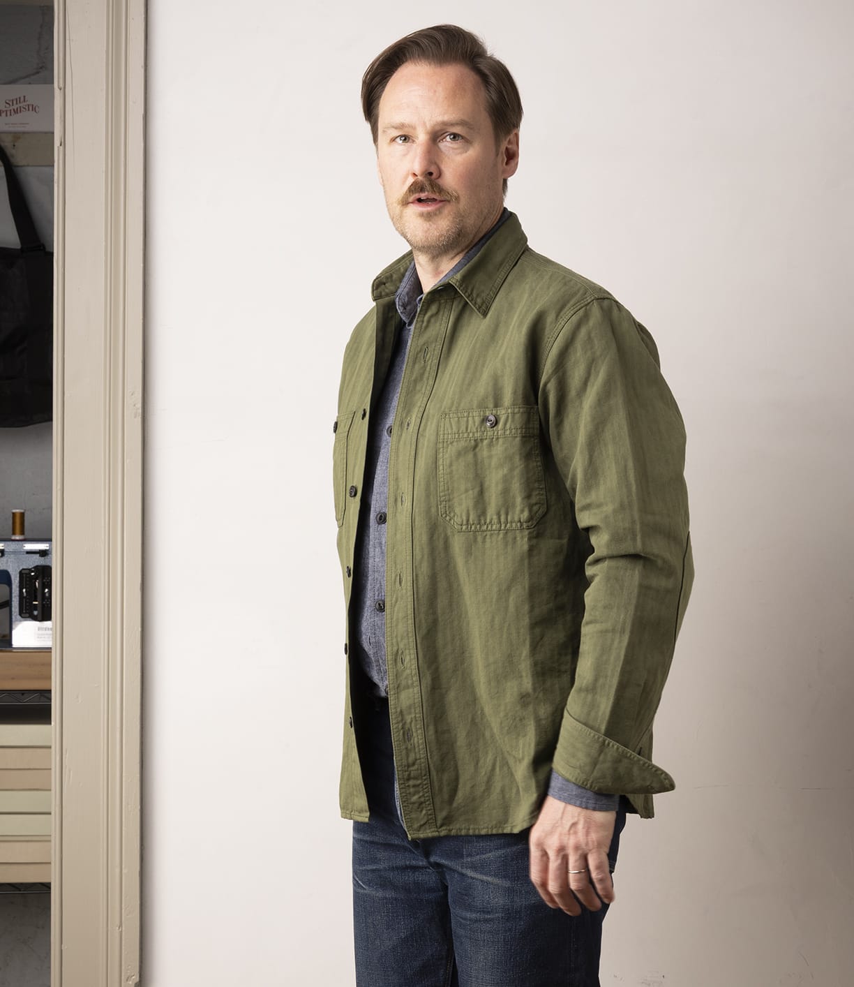A man with light brown hair and a mustache stands indoors, wearing the Best Made Co. Cotton-Linen Workshirt in Olive Drab over a gray shirt and blue jeans, looking slightly to his right against a plain light-colored wall.
