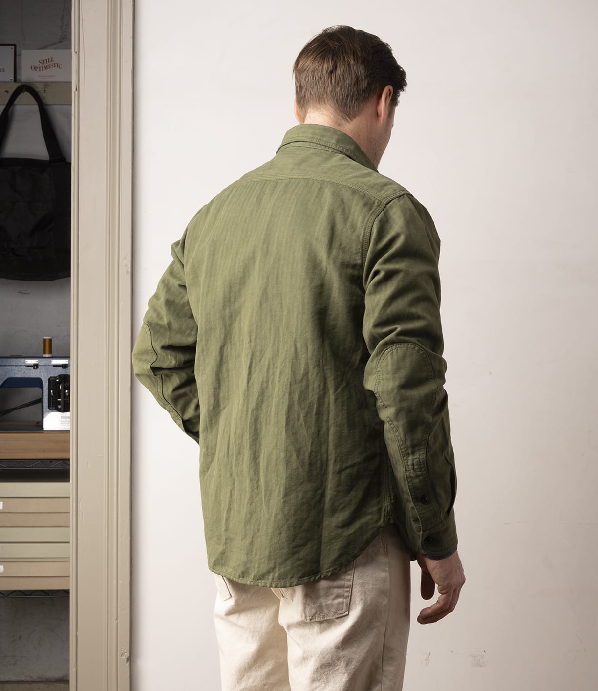 A person stands indoors facing away, wearing the Best Made Co. Cotton-Linen Workshirt in Olive Drab and beige pants. The creative room, with a sewing machine and drawers, evokes Japanese textile workwear inspiration.