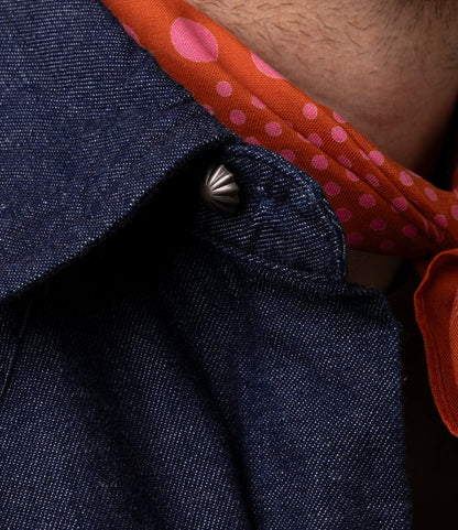 Close-up of a person wearing the Best Made Co. Fukuyama Snap-Front Shirt in Japanese indigo denim, styled with a metallic button and an orange scarf with pink polka dots. The edge of the person's beard is visible at the top.