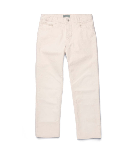 A light-skinned man in a blue shirt and Best Made Co Standard 5-Pocket Pant M2 Undyed stands indoors with hands behind his back. Behind him are a wooden chair and a shelf holding bags and books.