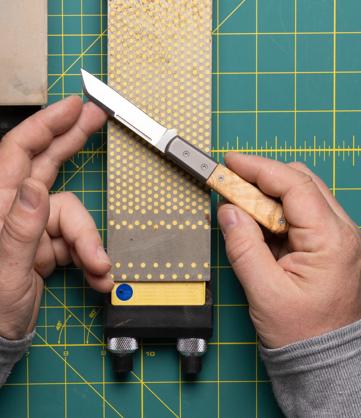A person holds a Best Made Co. Italian Barlow knife with an olive wood handle over a diamond sharpening stone on a green gridded cutting mat, gripping the knife in one hand and steadying the stone with the other.