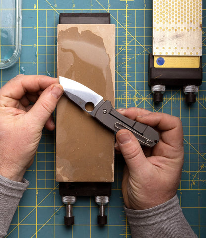 Someone sharpens a Best Made Co. Spyderco Techno 2 CTS-XHP pocket knife on a whetstone, both hands holding the blade at an angle on a green cutting mat, with sharpening tools and the titanium framelock nearby.