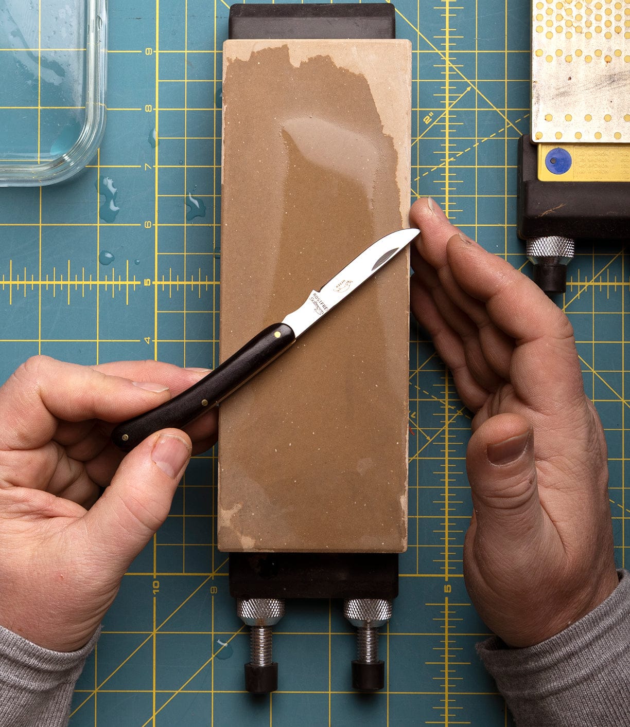 A person holds a Best Made Co Little Doctor Knife on a rectangular sharpening stone atop a cutting mat with measurement lines; another Otter-Messer sharpening tool is visible in the corner.