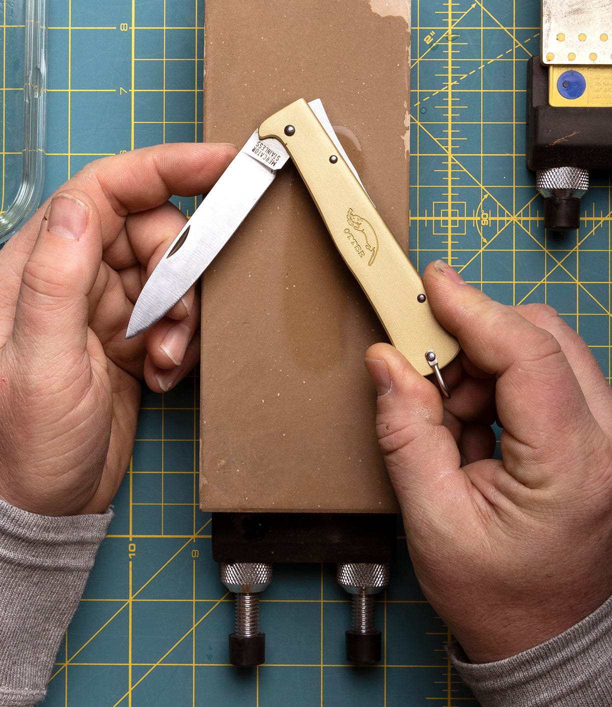 A person holds an open Best Made Co Brass Mercator pocket knife with a cream handle against a sharpening stone, placed on a green cutting mat with measurement lines and nearby tools visible.