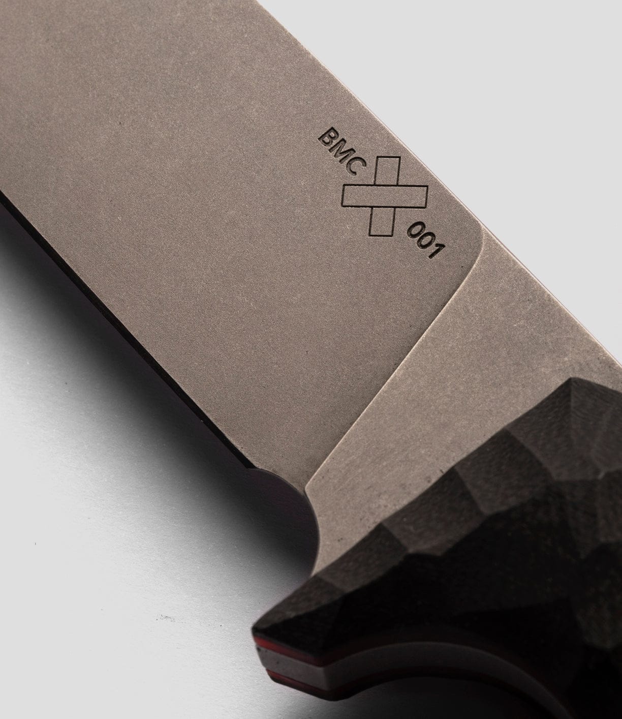 Close-up of The NWBW Field Knife (BMC Edition) by Best Made Co: metallic blade marked "BMC 001" with a cross, crafted from American D2 tool steel. A textured dark handle appears at right, set against a light gray background.
