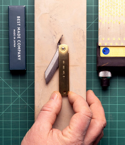A hand holds the Mid Brass Higo Knife by Best Made Co., its blade exposed on a sharpening stone. Beside it are a black Best Made Co. box and a yellow-capped sharpener, all arranged on a green cutting mat.