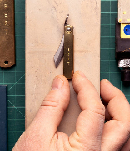 A hand holds the Mini Brass Higo Knife by Best Made Co., engraved with "BEST," resting on a beige rectangular surface atop a green grid-lined cutting mat, inspired by Japanese folding knives and handmade Edo-period design.